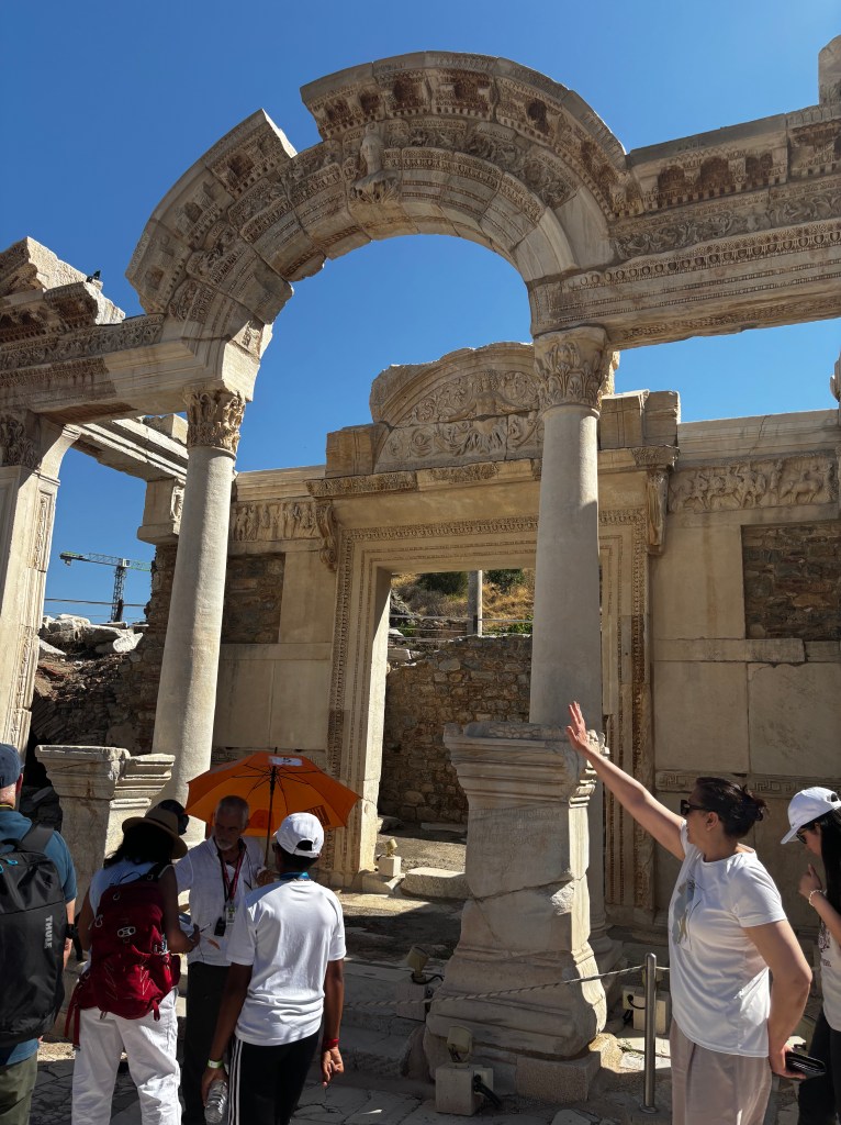 A historic archway in Ephesus, surrounded by tourists, with a tour guide speaking and an orange umbrella in the background.